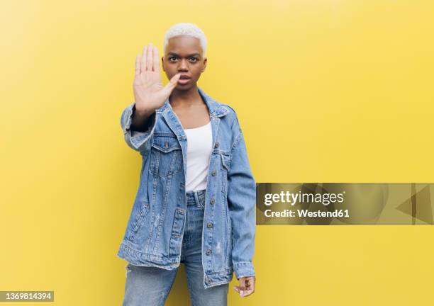 serious woman making stop gesture standing against yellow background - hand geste halt stock-fotos und bilder