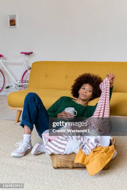 bored woman holding t-shirt and coffee mug reclining on sofa at home - bored housewife stock-fotos und bilder