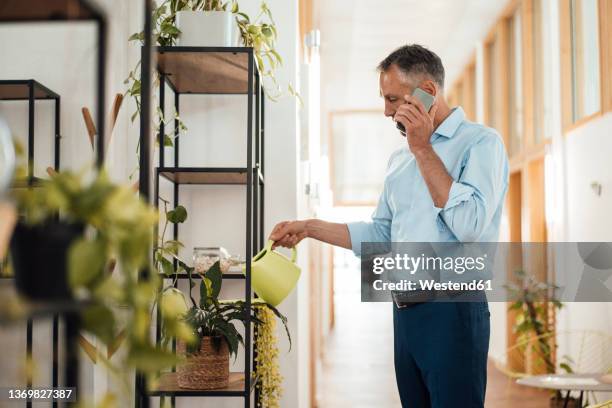 businessman talking on mobile phone watering plant at corridor in office - watering plants stock-fotos und bilder