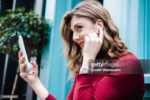 smiling woman with smart phone adjusting bluetooth earphones - ohrhörer stock-fotos und bilder