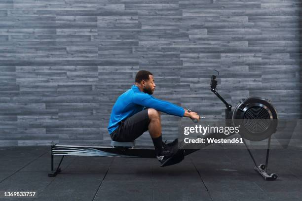 concentrated athlete exercising on rowing machine in front of wall in gym - rowing machine stock pictures, royalty-free photos & images