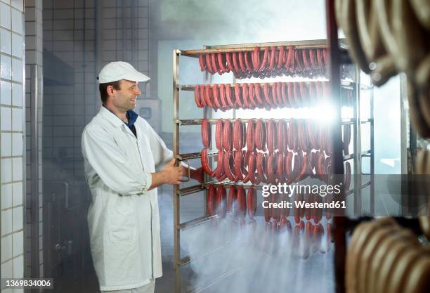 smiling butcher looking at arranged sausages in smokehouse - smokehouse stock pictures, royalty-free photos & images