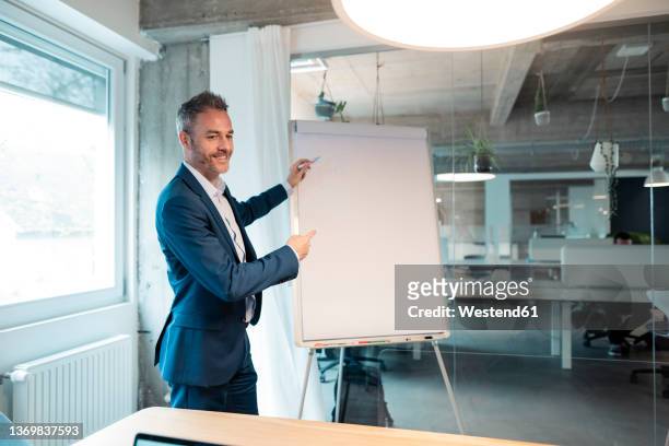smiling businessman writing on flipchart gesturing at office - dreiviertelansicht stock-fotos und bilder