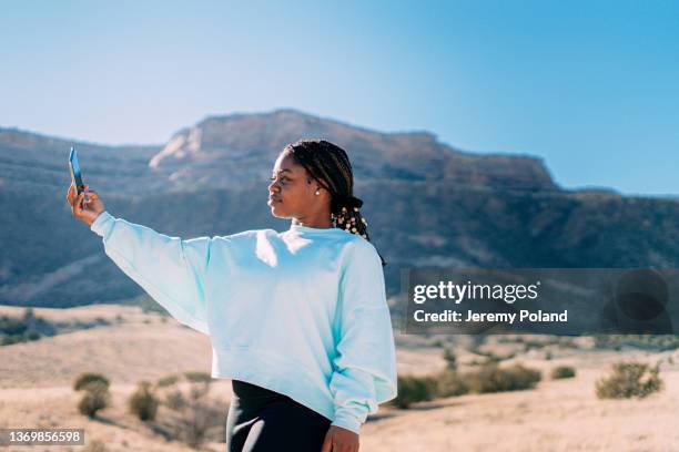 cheerful young african american woman stopping to take a selfie or vlog on her smartphone walking on a footpath outdoors - american influencer stock pictures, royalty-free photos & images