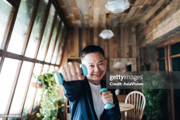 active senior asian man exercising at home. practicing fitness training and lifting dumbbells. looking at camera with smile. maintaining healthy fitness habits. elderly wellbeing, health and wellness concepts - elderly cognitive stimulation therapy stockfoto's en -beelden