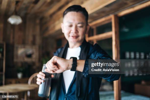 active senior asian man resting after exercising at home, looking at his smartwatch, using fitness tracker app and measuring pulse. maintaining healthy fitness habits. elderly wellbeing, health, wellness and technology concept - man doing yoga in the morning stockfoto's en -beelden