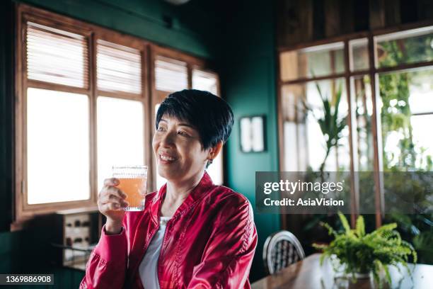 active senior asian woman in sportswear drinking a glass of pink fruit juice after exercising at home, staying hydrated while taking a break from daily training - juice drink stock pictures, royalty-free photos & images