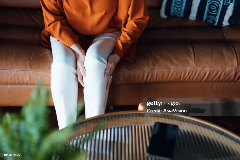 Abgeschnittene Aufnahme einer verzweifelten älteren asiatischen Frau, die sich unwohl fühlt und unter Schmerzen im Bein leidet, während sie zu Hause auf dem Sofa im Wohnzimmer sitzt. Konzept für ältere Menschen und Gesundheitsfragen