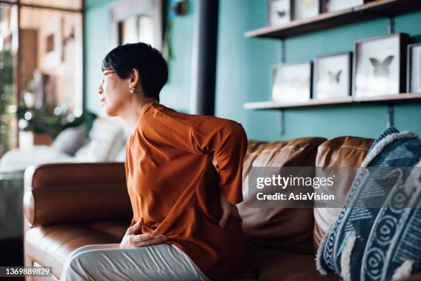 取り乱したアジアの女性は、腰痛に苦しみ、自宅のリビングルームのソファに座っている間に筋肉をマッサージし、気分が悪い。高齢者と健康問題の概念 - 人の筋肉 ストックフォトと画像