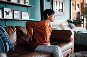 A distraught senior Asian woman feeling unwell, suffering from backache, massaging aching muscles while sitting on sofa in the living room at home. Elderly and health issues concept
