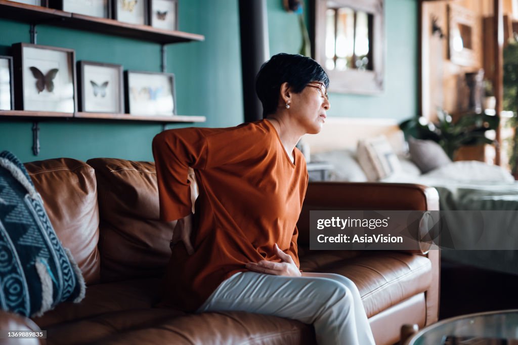 A distraught senior Asian woman feeling unwell, suffering from backache, massaging aching muscles while sitting on sofa in the living room at home. Elderly and health issues concept