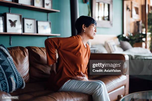 a distraught senior asian woman feeling unwell, suffering from backache, massaging aching muscles while sitting on sofa in the living room at home. elderly and health issues concept - 下背部痛 個照片及圖片檔