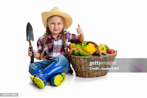 Portrait of cute little girl, emotive kid in image of farmer, gardener sitting near giant basket of vegetables isolated on white background. Concept of job, work, childhood, games