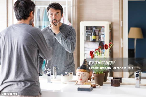 handsome bearded 50 years old man looking at himself in bathroom mirror at home. self care concept. - 50 54 years stock-fotos und bilder