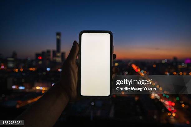 a hand holding a mobile phone with a white screen with a city in the background - hand holding iphone stock pictures, royalty-free photos & images