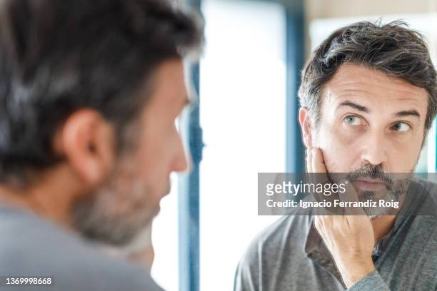 portrait of a handsome bearded 50 years old man looking in the mirror while  touching his beard in bathroom at home. self care concept. - 50 years old man imagens e fotografias de stock