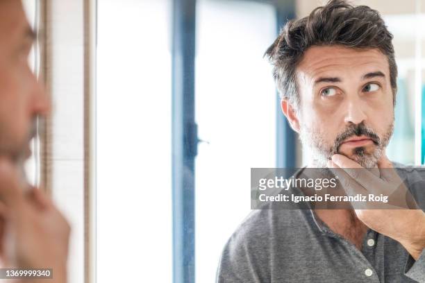 portrait of a handsome bearded 50 years old man looking in the mirror while touching his beard in bathroom at home. self care concept. - 50 54 years stock-fotos und bilder
