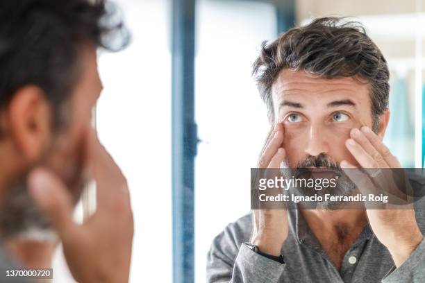 portrait of a handsome bearded 50 years old man looking at his wrinkles  in  the bathroom mirror at home. self care concept. - 50 54 years stockfoto's en -beelden