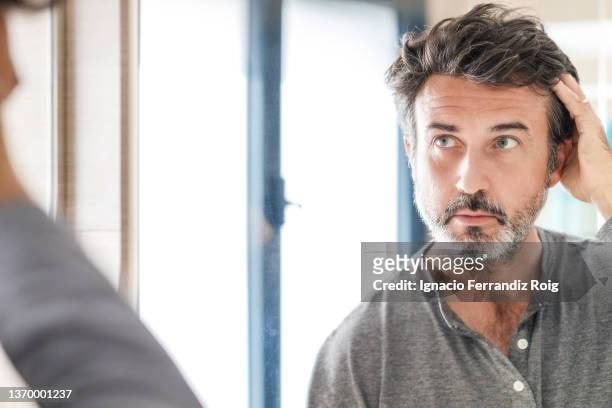 portrait of a handsome bearded 50 years old man looking at his white hair  in  the bathroom mirror at home. self care concept. - homme coiffure photos et images de collection