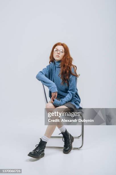 young red-haired teenage girl in glasses sits on a chair in the studio and poses. - sitting in a chair stockfoto's en -beelden