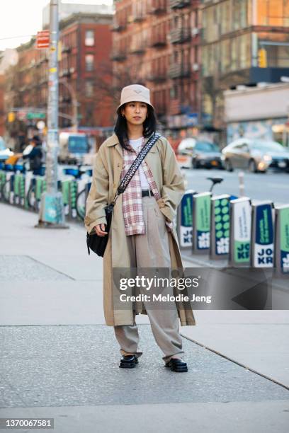 Kristen Lam of Moda Operandi wears a tan Prada bucket hat, tan trench coat, white top, red print shirt, black cross-body bag, tan slouchy pants, and...