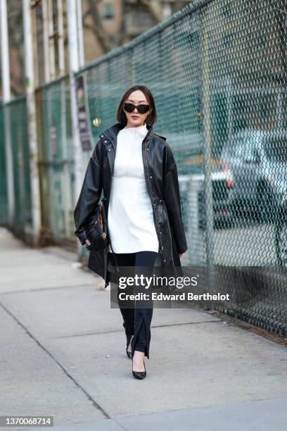 Chriselle Lim wears sunglasses, a long oversized leather jacket, a white short dress worn as a top, a black bag with leather fringes, black slit...