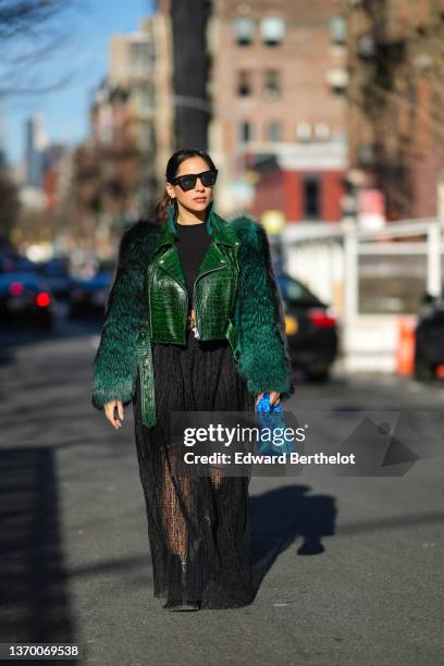 Samia Laaboudi wears sunglasses, green long fringed earrings, a black top, a green leather jacket with fluffy faux fur sleeves, a black ribbed top, a...