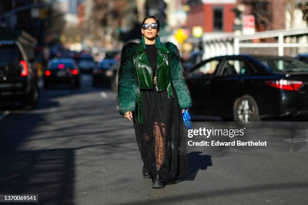 Samia Laaboudi wears sunglasses, green long fringed earrings, a black top, a green leather jacket with fluffy faux fur sleeves, a black ribbed top, a...