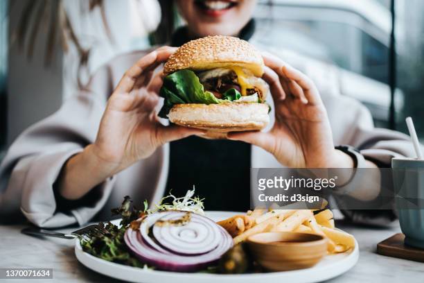 close up of happy young asian woman sitting by the window in a restaurant, enjoying lunch during the day. she is having freshly made delicious burger with fries and side salad. lifestyle, people and food concept - woman eating burger stock pictures, royalty-free photos & images