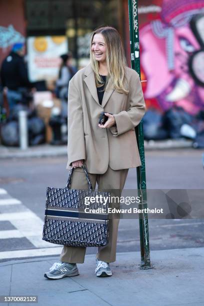 Guest wears a black t-shirt, an oversized beige blazer jacket, beige large suit pants, a beige and navy blue Dior Oblique Jacquard large Book-Tote...