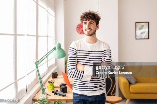 young man standing with crossed arms in his modern studio while looking away - casual male stockfoto's en -beelden