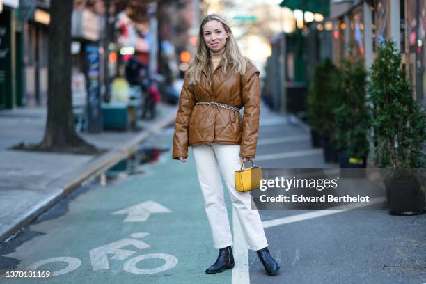 Guest wears a brown / beige ribbed turtleneck pullover, earrings, a brown shiny leather quilted leather puffer jacket, silver chains belt, white...