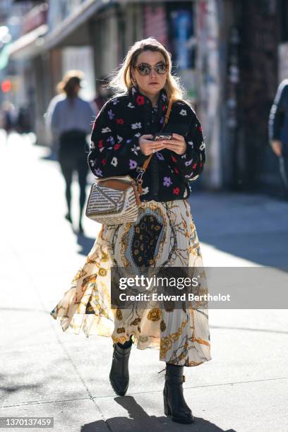 Guest wears brown sunglasses, gold earrings, a brown and beige print pattern shirt, a black with embroidered red / white / purple fluffy flower...