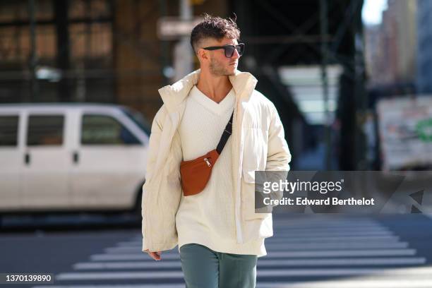 Guest wears sunglasses, a white oversized V-neck pullover, a white high neck puffer jacket, a brown fanny pack bag, gray pants, outside New York...
