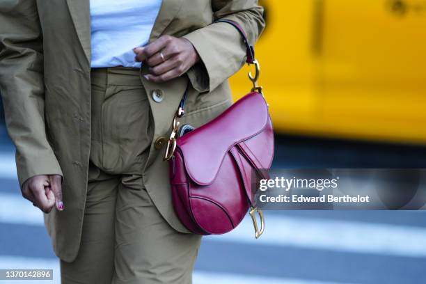 Guest wears a white t-shirt, a brown blazer jacket, matching brown large pants, a burgundy shiny leather Saddle handbag from Dior, gold rings,...