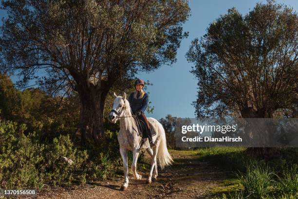 young smiling blond woman during her horseback ride in the mallorcan countryside between olive trees - riding habit stock pictures, royalty-free photos & images