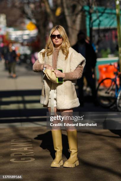 Guest wears sunglasses, a brown fluffy pullover, a beige shiny leather with large sheep borders sleeveless oversized jacket, white leather shorts, a...