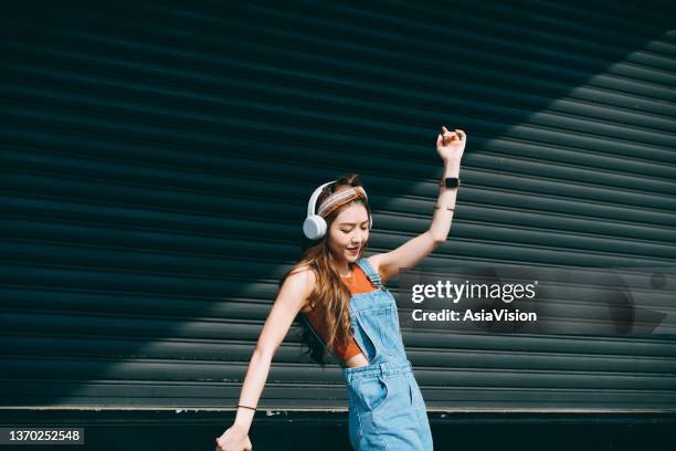 carefree, smiling young asian woman dancing with her eyes closed while listening to music on headphones outdoors against coloured wall and sunlight. music and lifestyle - skaka på huvudet bildbanksfoton och bilder