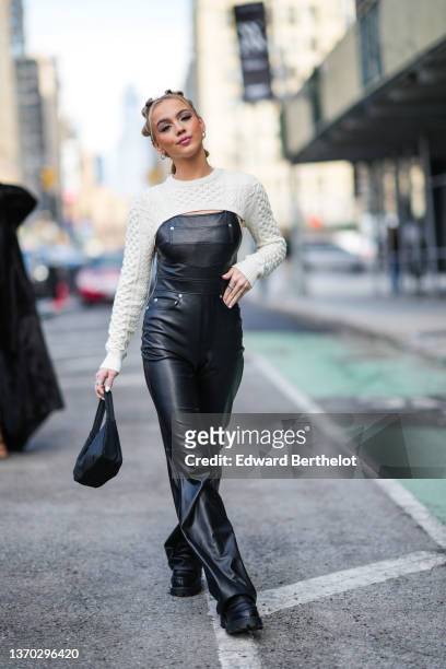 Guest wears silver earrings, a white braided cool cropped pullover, a black shiny leather pair of overalls, silver rings, a black nylon Reedition...