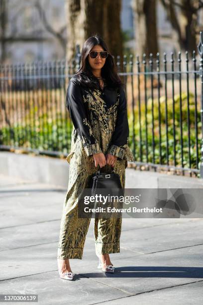 Guest wears brown sunglasses, gold chain necklaces, a black with yellow print pattern satin shirt, matching black with gold print pattern satin...