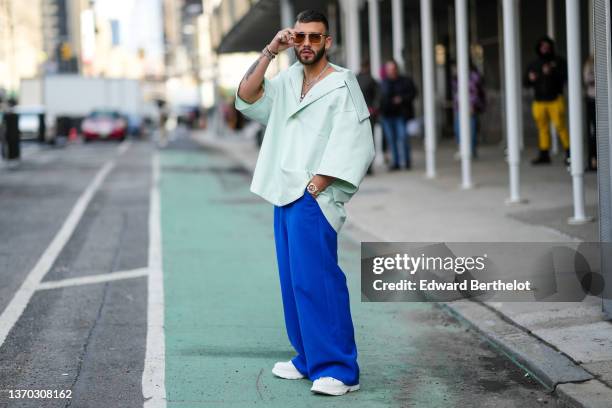 Guest wears sunglasses, silver and black monogram earrings from Louis Vuitton, silver chain and rhinestones necklaces, a pale green oversized short...