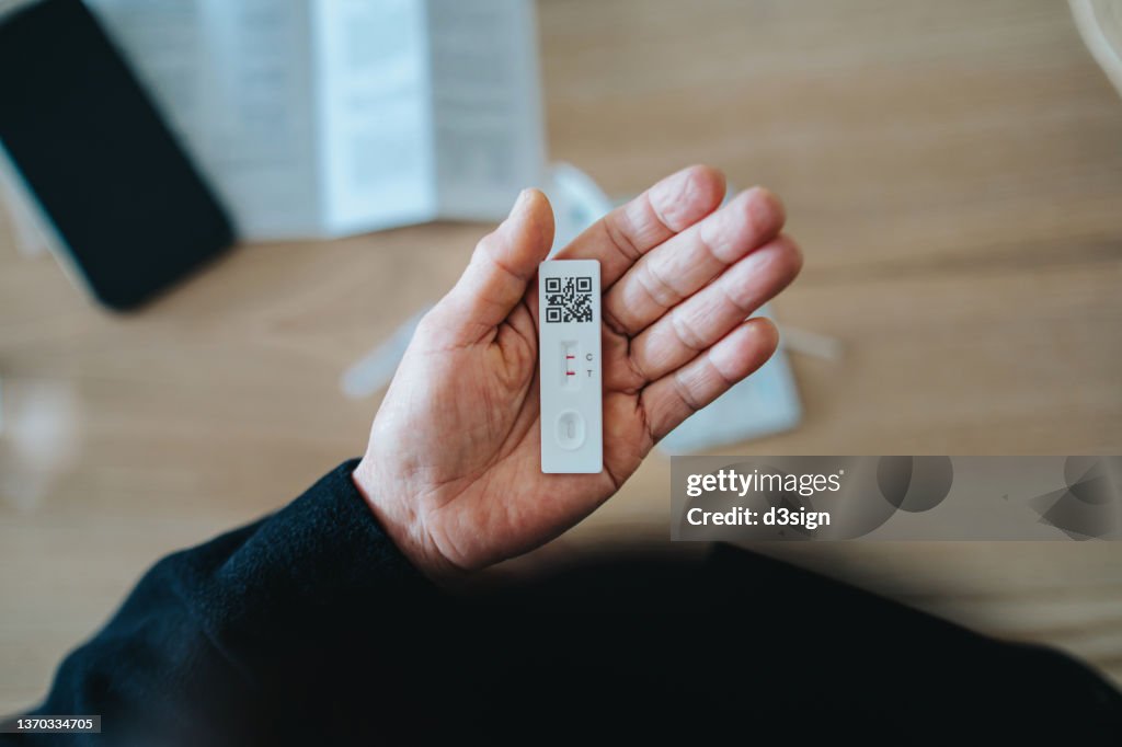 Overhead view of senior Asian woman carrying out a Covid-19 rapid lateral flow test at home. She is holding a positive Coronavirus rapid self test device, feeling worried