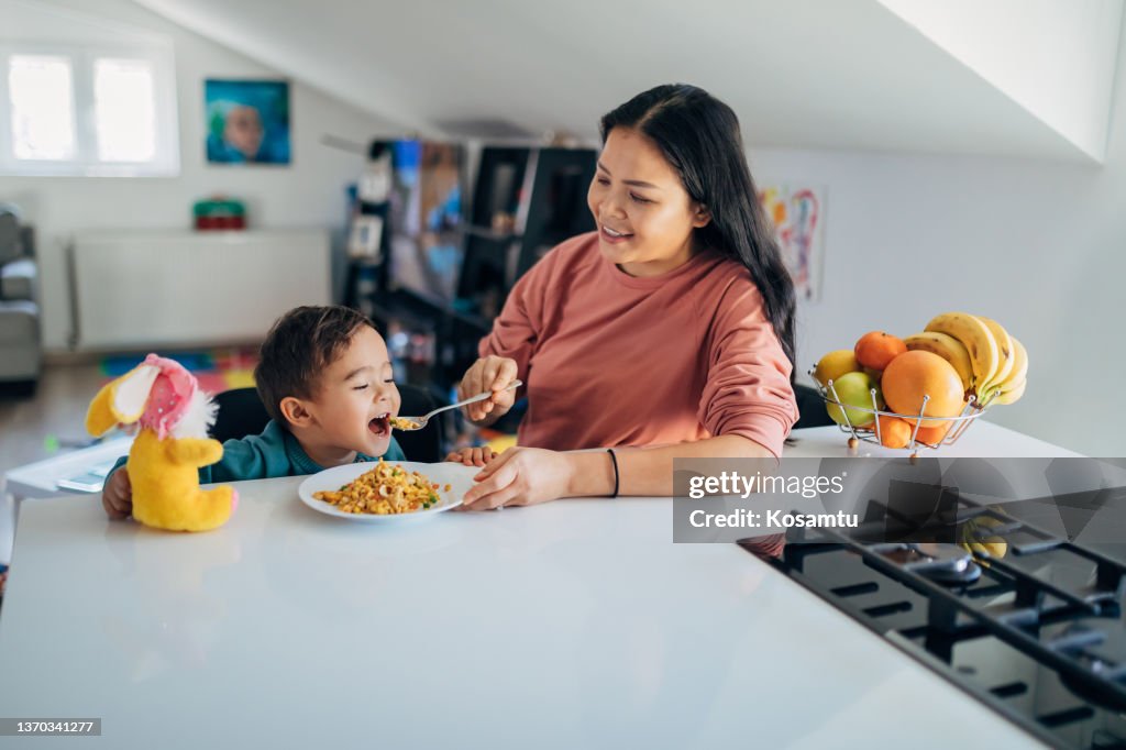 Eine fürsorgliche und lächelnde Mutter füttert ihren süßen kleinen Sohn. Sie sitzen am Esstisch.