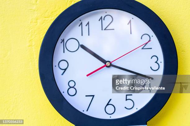 closeup of a white wall clock with black numbers hanging on a yellow wall. - wall clock stock pictures, royalty-free photos & images