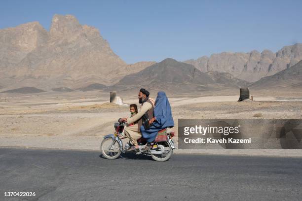 Pashtun man with his wife in a blue burka in the back and his daughter sat in the front rides a motorbike on the outskirts of the district on...