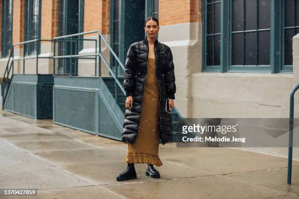 TyLynn Nguyen wears a long black Khaite down jacket, light brown dress, and black boots at the Khaite show on February 13, 2022 in New York City.