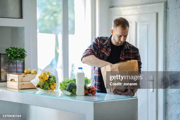 young man taking groceries out of paper bag on the counter at home - young man groceries kitchen stock pictures, royalty-free photos & images