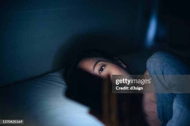 young asian woman surfing on the net on smartphone while lying in bed at cozy bedroom in late night. woman with obscured face using mobile phone, device screen light illuminated on her. lifestyle, habits and technology - insomnia stock pictures, royalty-free photos & images
