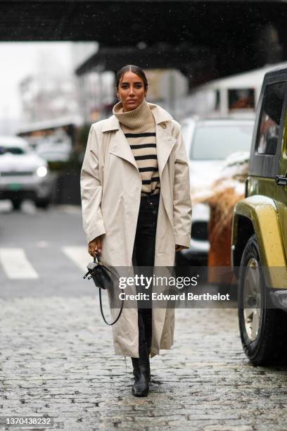 Tamara Kalinic wears gold earrings, a beige and black striped print pattern turtleneck pullover, a beige long trench coat, black denim jeans pants, a...