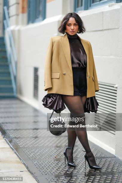 Kat Graham wears gold large earrings, a black high neck satin blouse with oversized puffy ruffled sleeves, a brown beige oversized blazer jacket,...